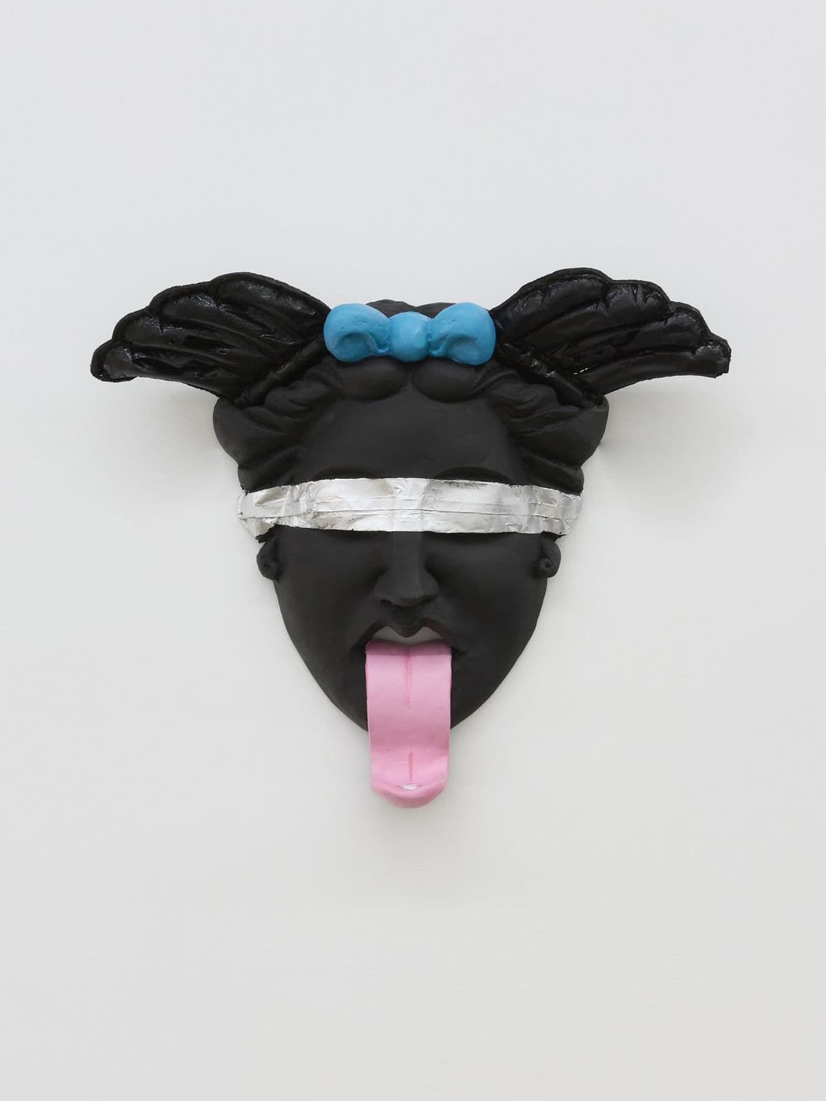 Oral Tradition II, black mythological mask sculpture by Grigorios Ilaridis with wing-like forms, a silver band across the eyes and a coin resting on an extended tongue.